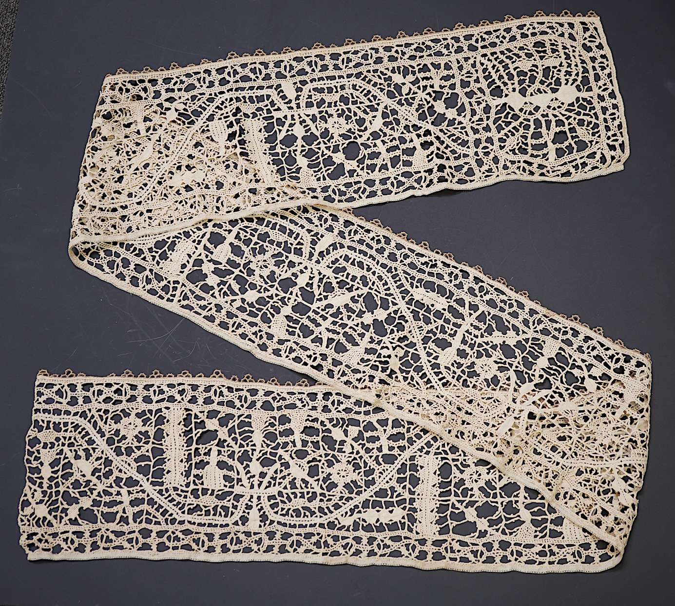 A length of late 16th early 17th century Venetian Reticella needle lace. Possibly cut from a wider panel or the edge to an Ecclesiastical robe or a furnishing lace. Designed with figures, worked in button hole stitch. 17